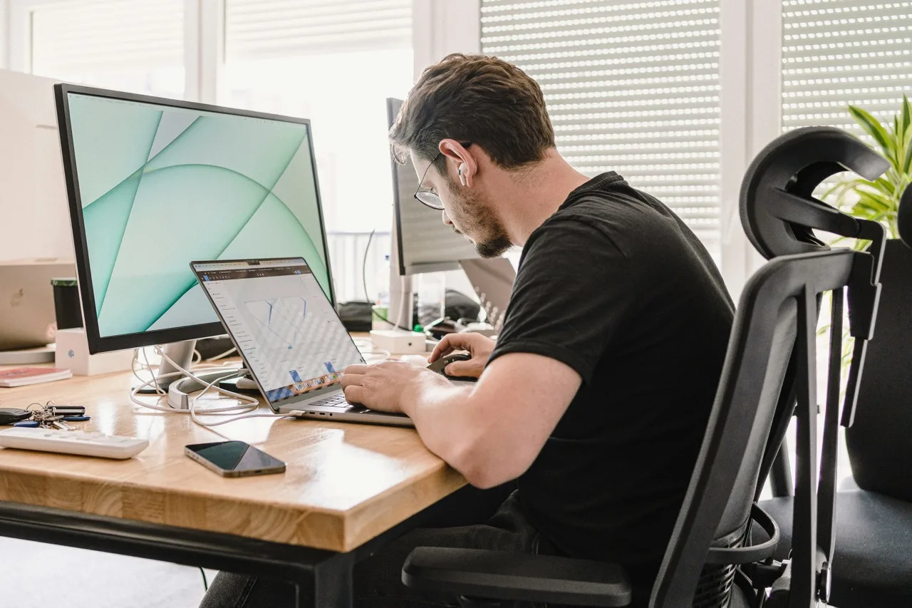 A person working on a wooden desk, hes looking at a laptop but there is also a large monitor behind it. The man has dark hair and is wearing a black t shirt sat on a black office chair