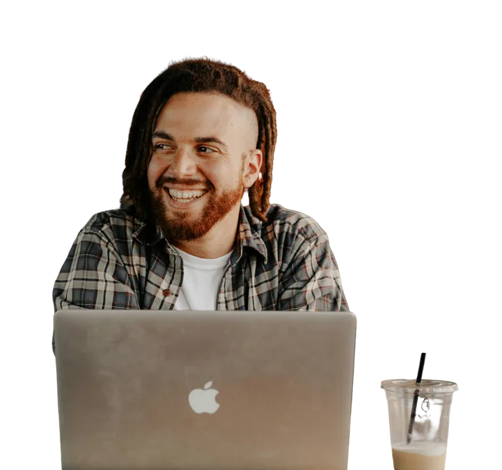 Man with ginger long hair and a beard smiling looking away from the camrea in front of a macbook and with a smoothie next to him.