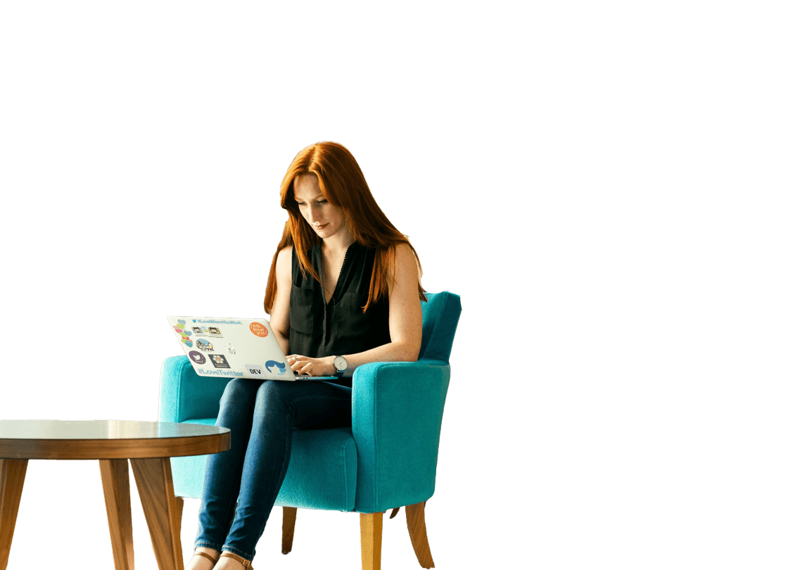 Women working on chair with laptop on her knees, she has ginger hair and is sat next to a wooden table.