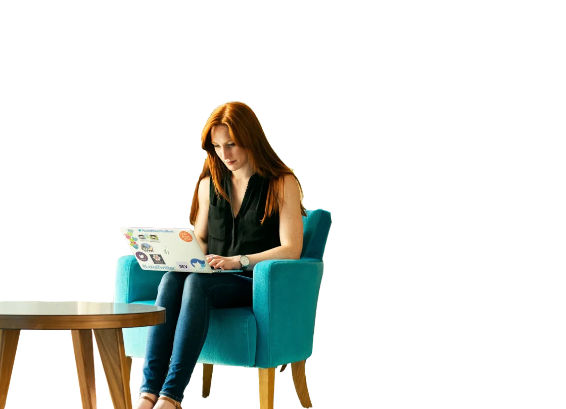 Women working on chair with laptop on her knees, she has ginger hair and is sat next to a wooden table.
