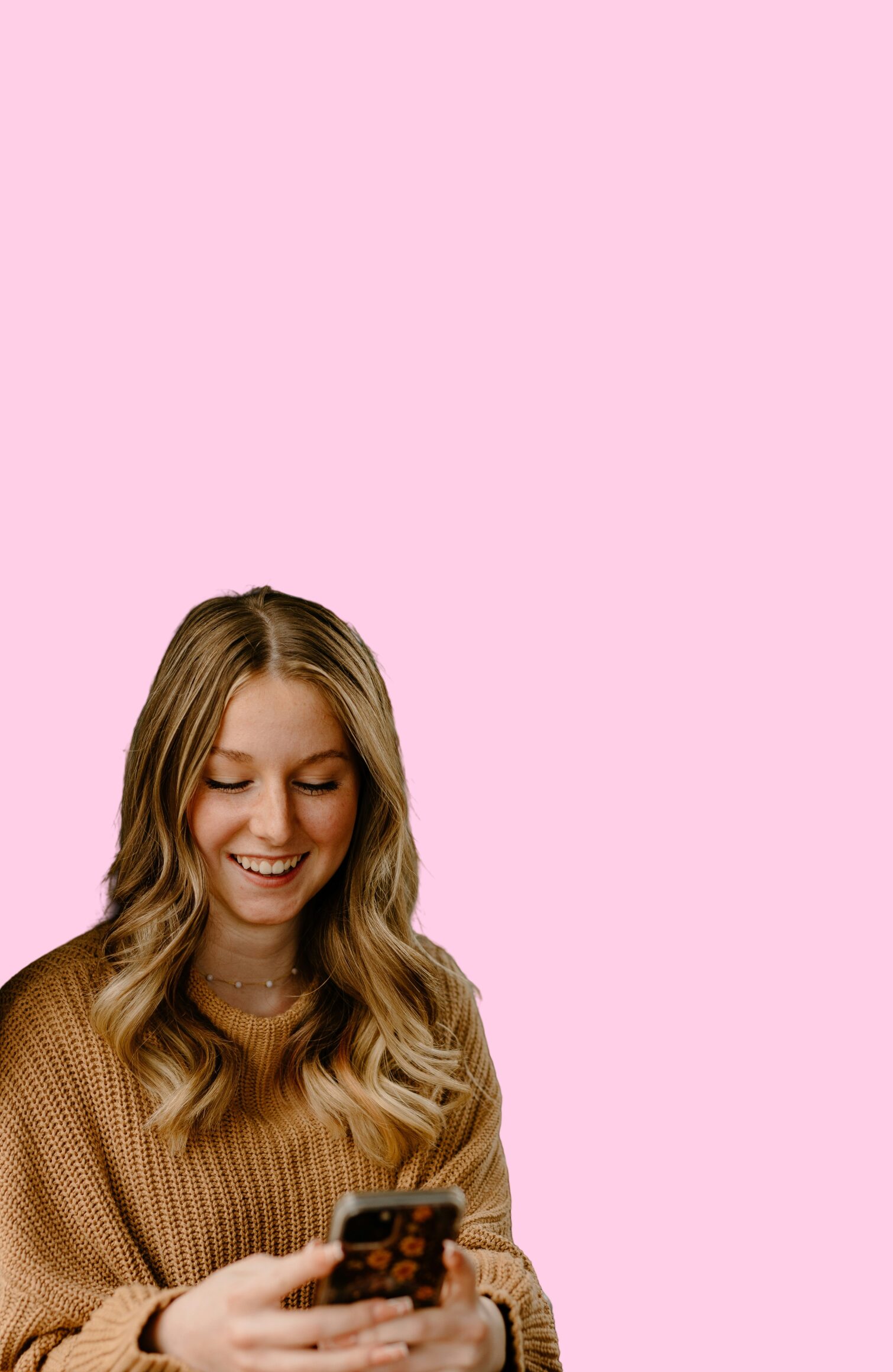 A young woman smiling while looking at her phone, set against a pink background.