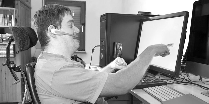 Person using an adaptive computer setup with a headset and touchscreen monitor at a desk.
