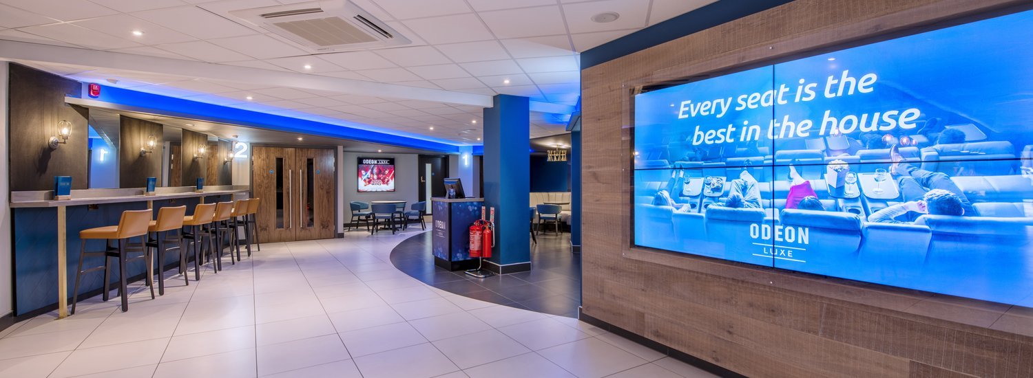 Odeon Luxe cinema foyer with blue illuminated wall display, seating area, and corridor leading to screening rooms.