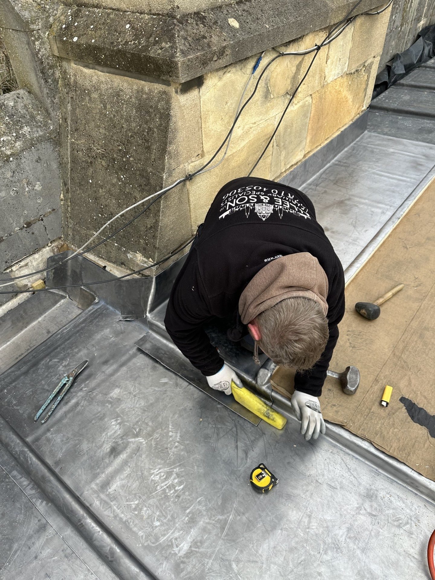 A roofer in an N Lee and Son jacket kneeling on a lead roof, dressing a lead sheet by hand against a stone parapet, with a mallet, dresser and hand tools laid out beside him